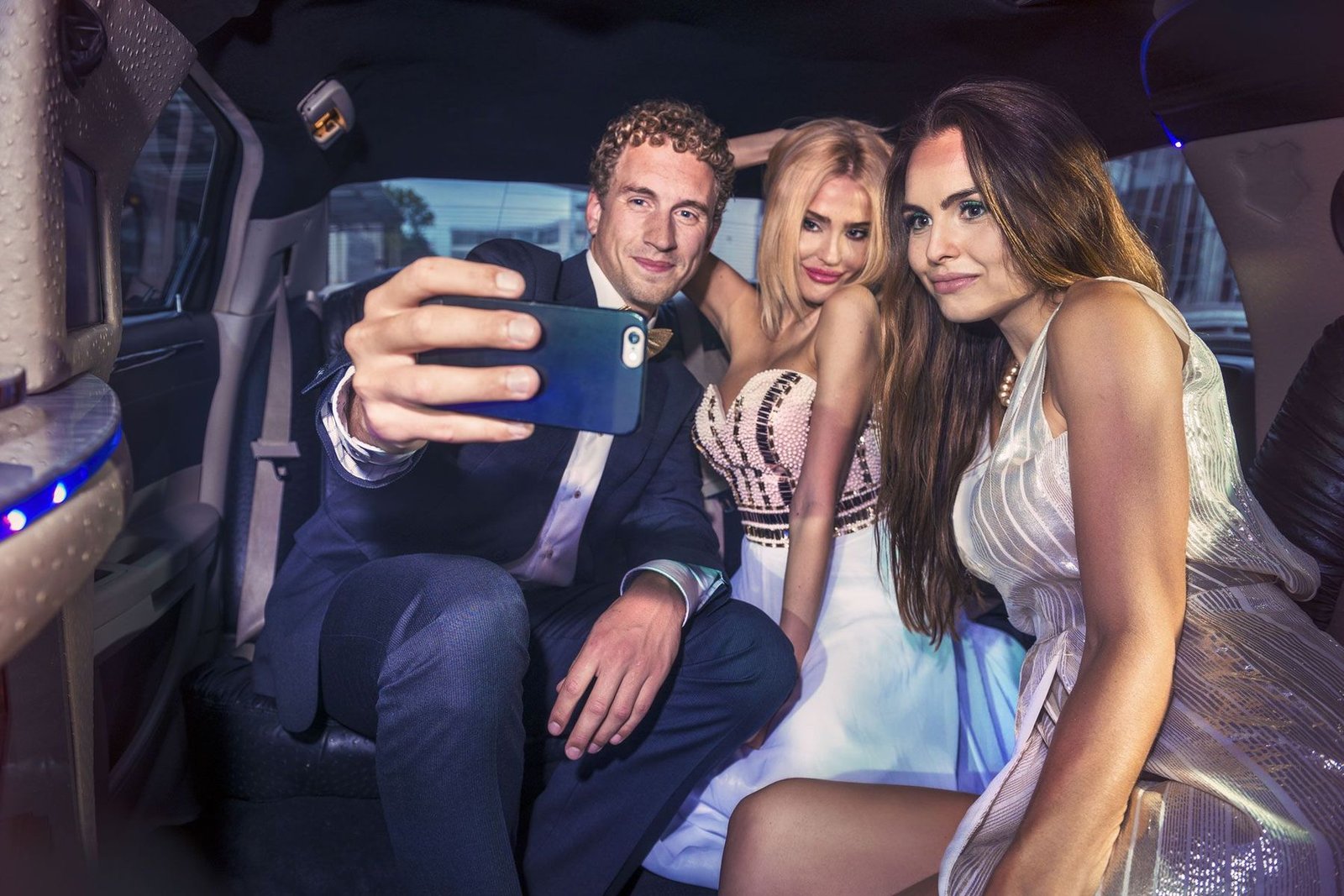 A person in a suit takes a selfie with two people in formal wear inside a limousine